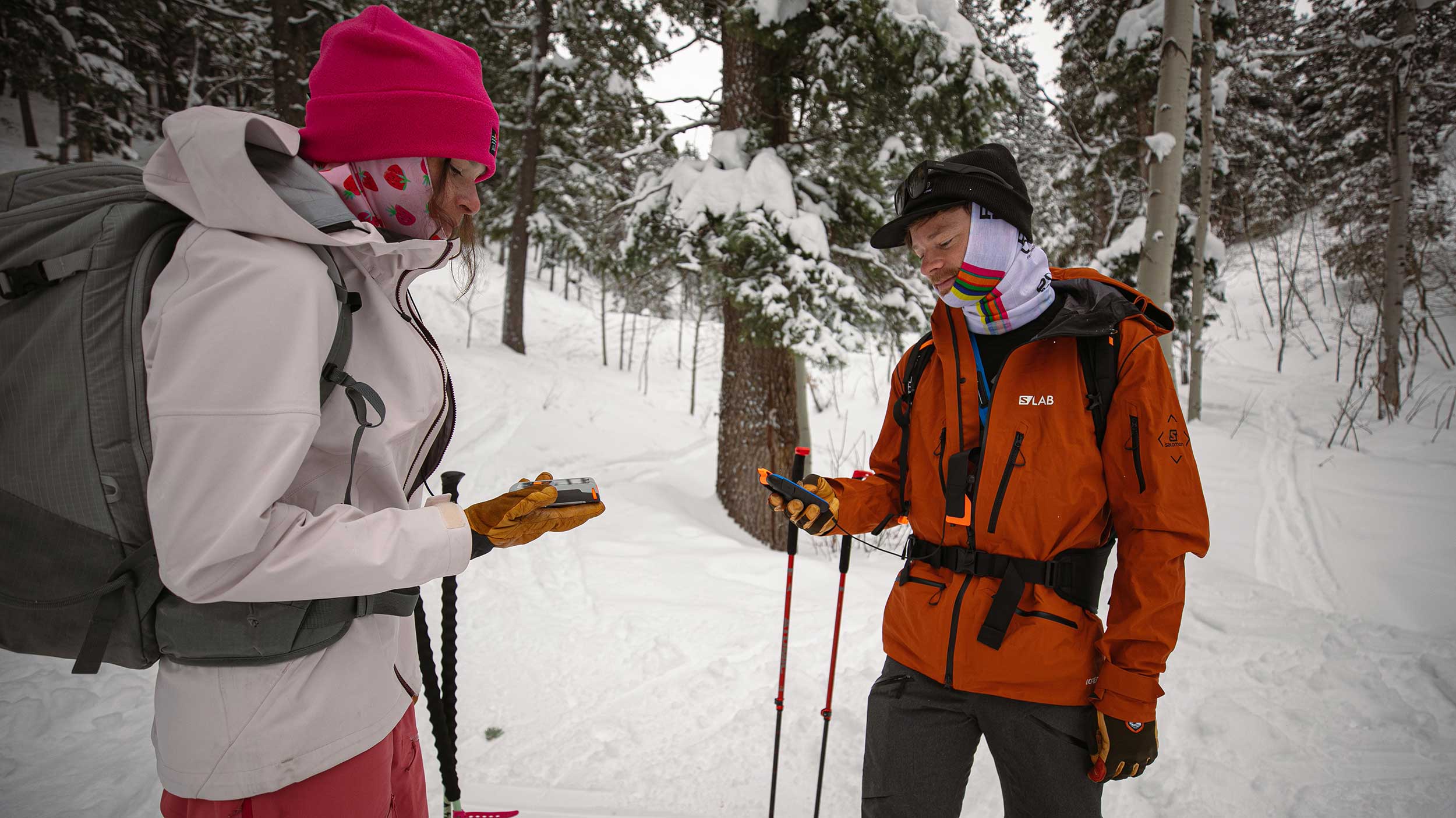Two people in winter gear standing in a snowy forest with backpacks and ski poles.