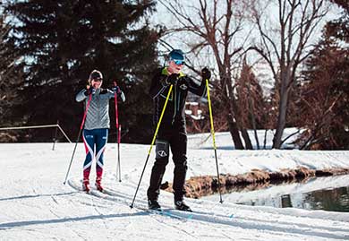 Two people cross-country skiing on a snow-covered path with trees and a pond in the background.