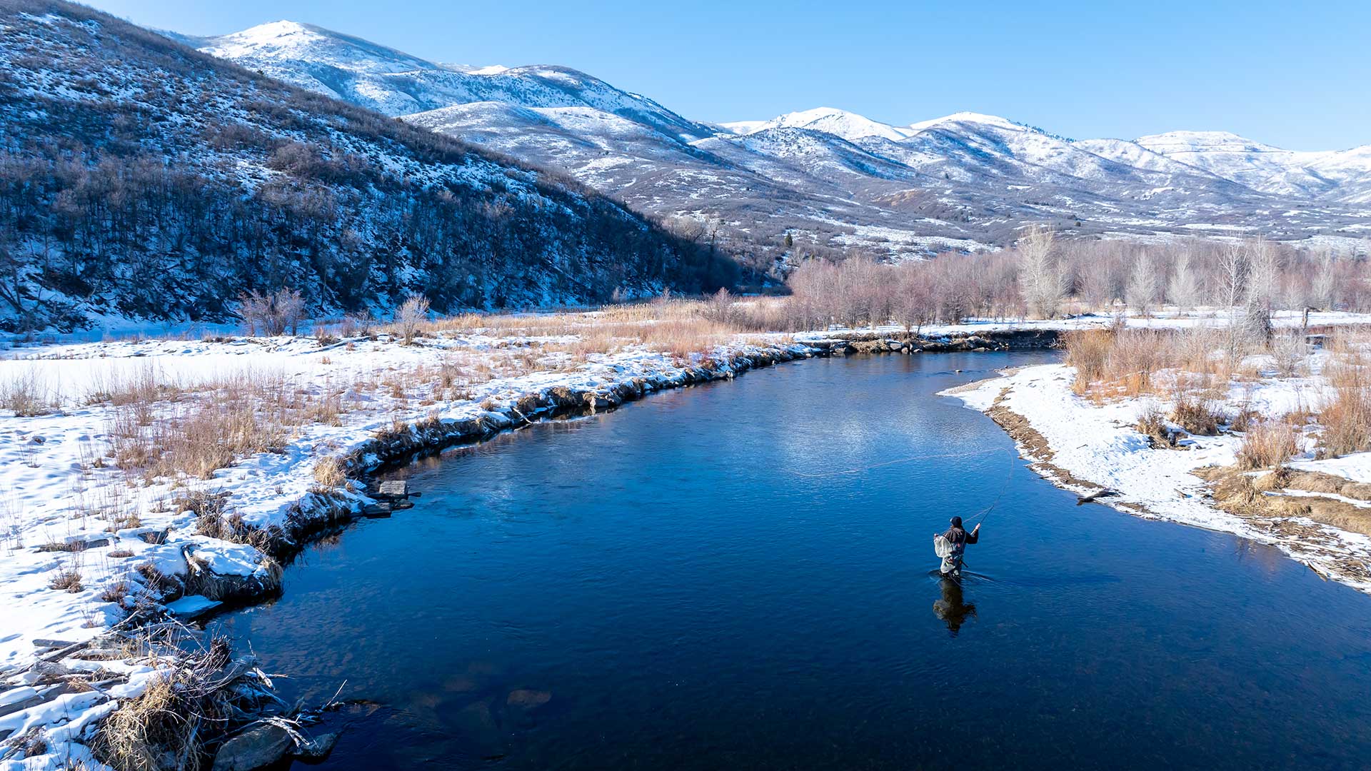 Person fly fishing in a river with snow-covered mountains in the background