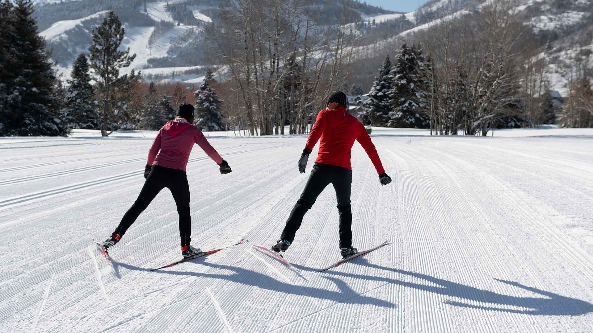Two people cross-country skiing on a snow-covered track with mountains in the background