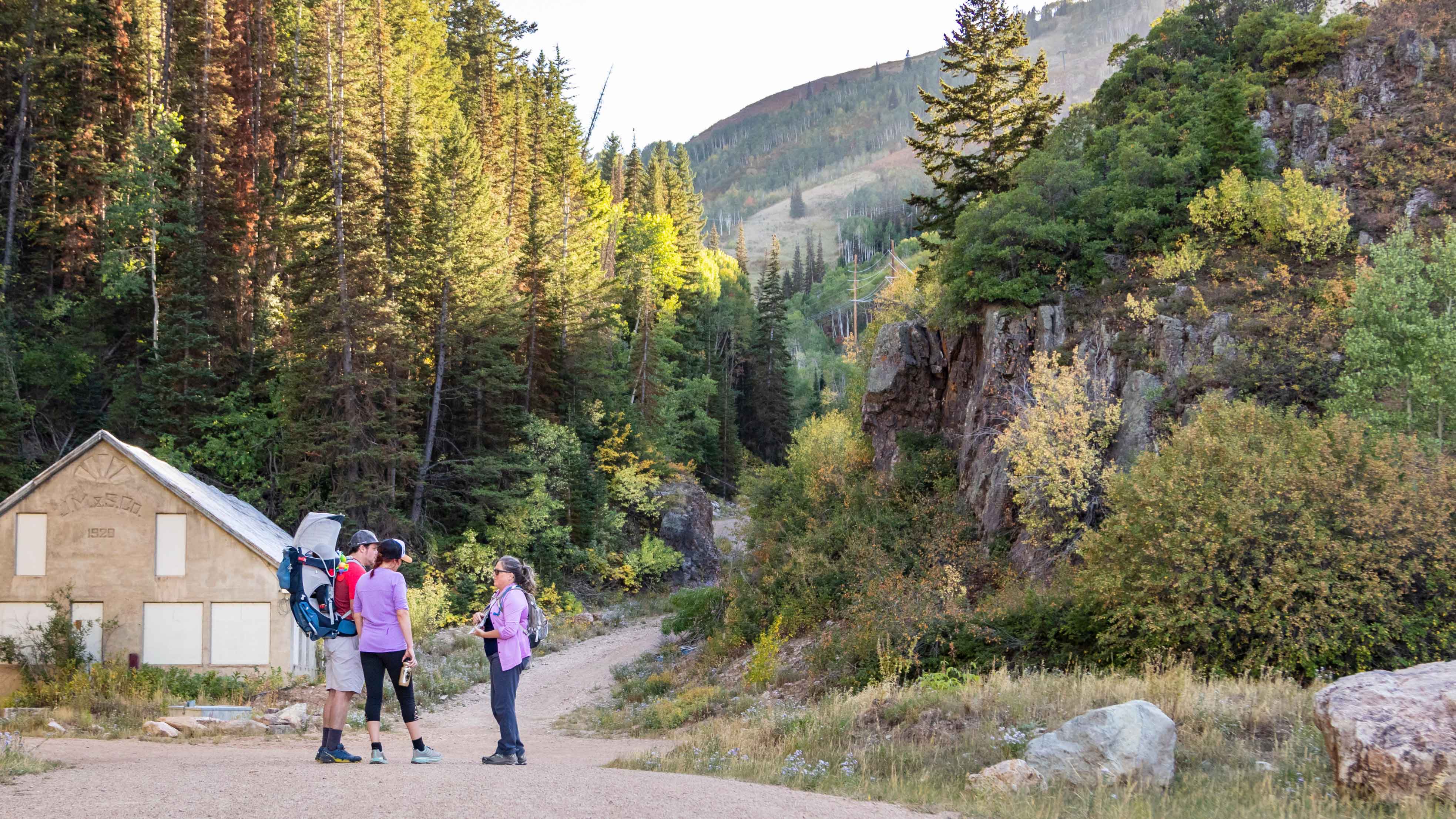 Group of people hiking on a trail in a forested area with a wooden building in the background.