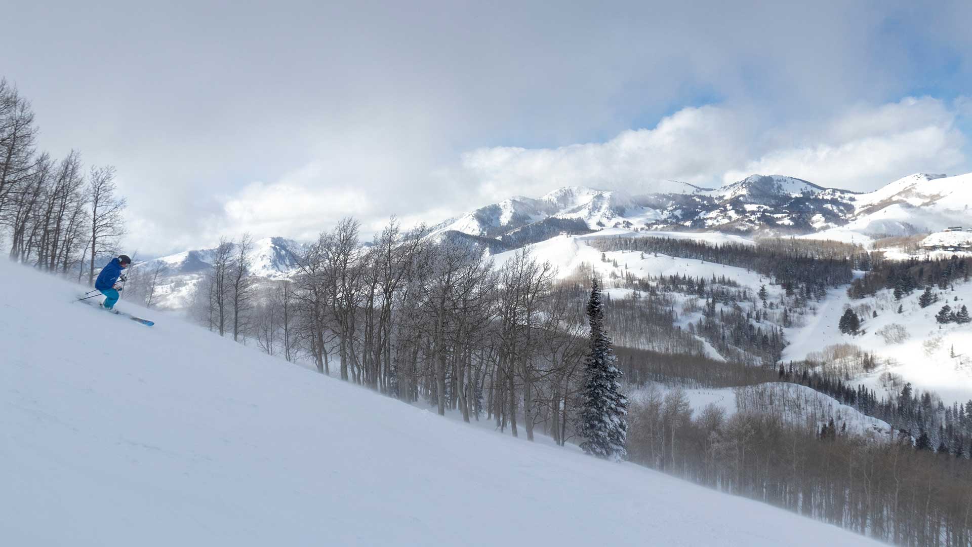 Jans Expert skiing in a blue outfit down a snowy mountain slope with bare trees in the foreground and snow-covered mountains under a cloudy blue sky in the background while skiing at Deer Valley Resort, Park City, Utah.