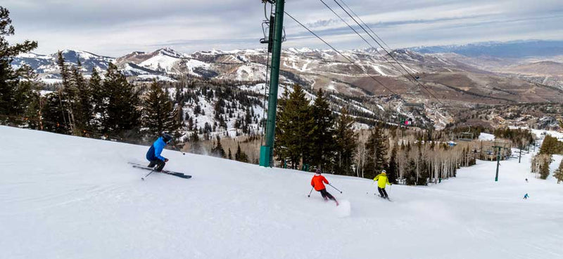 Three skiers on a snowy slope with a ski lift and mountainous landscape in the background.