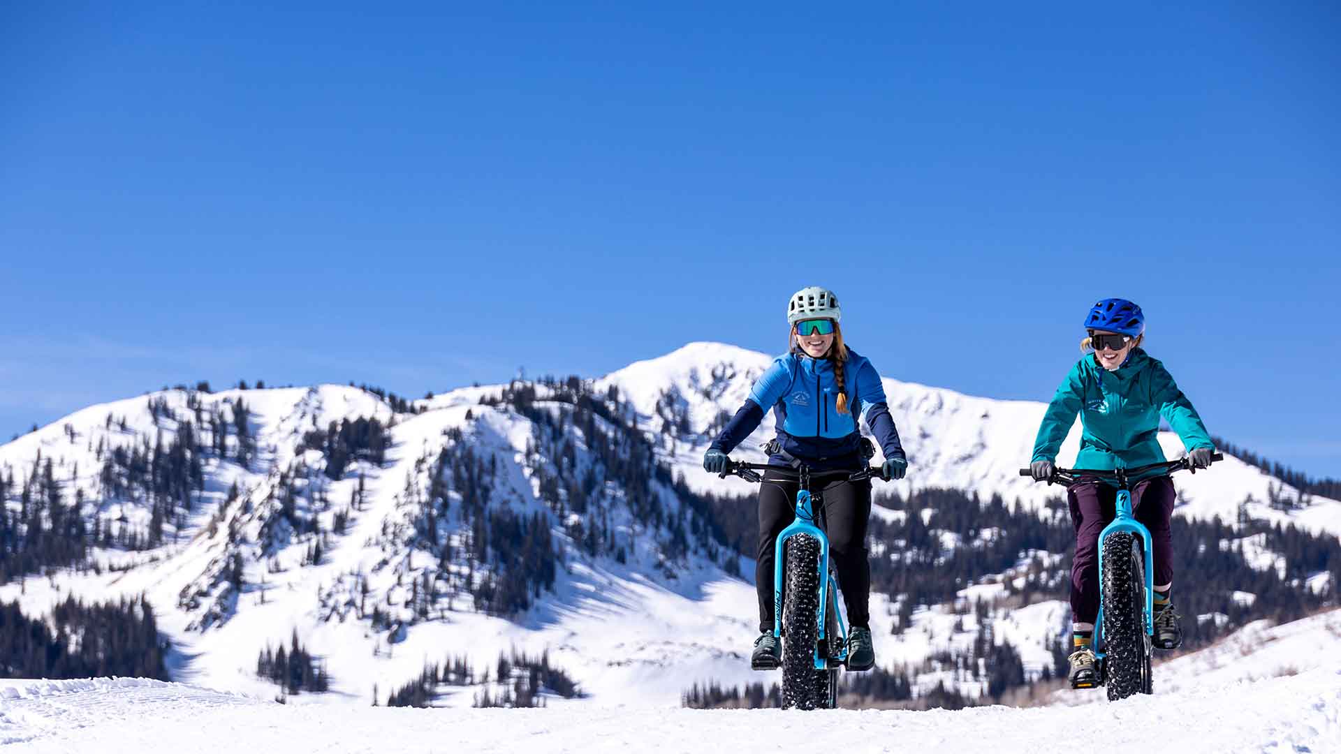 Two people riding fat-tire bikes on a snowy landscape with mountains in the background.