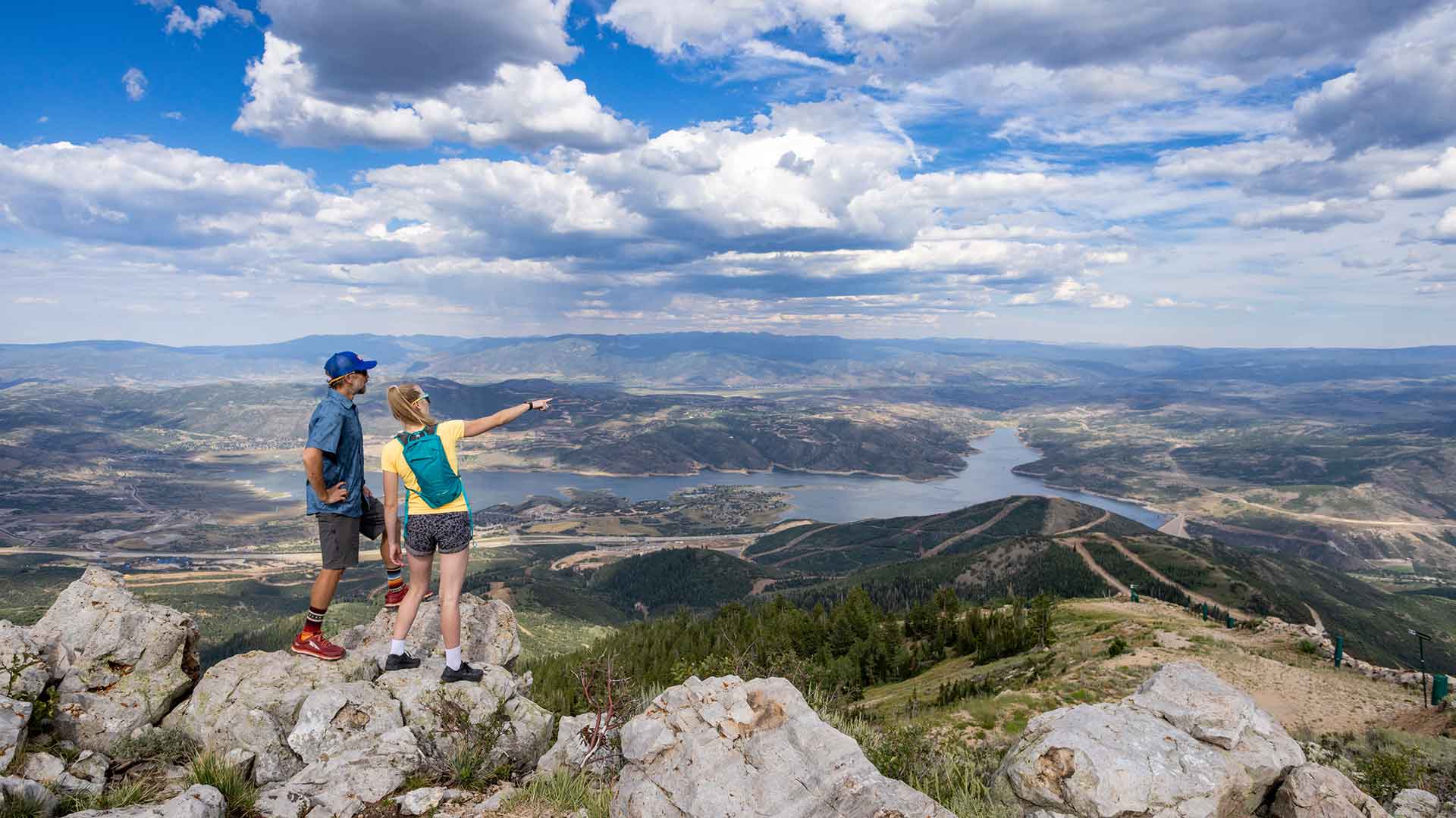 Two people standing on a rocky outcrop overlooking a scenic landscape with a lake and mountains.
