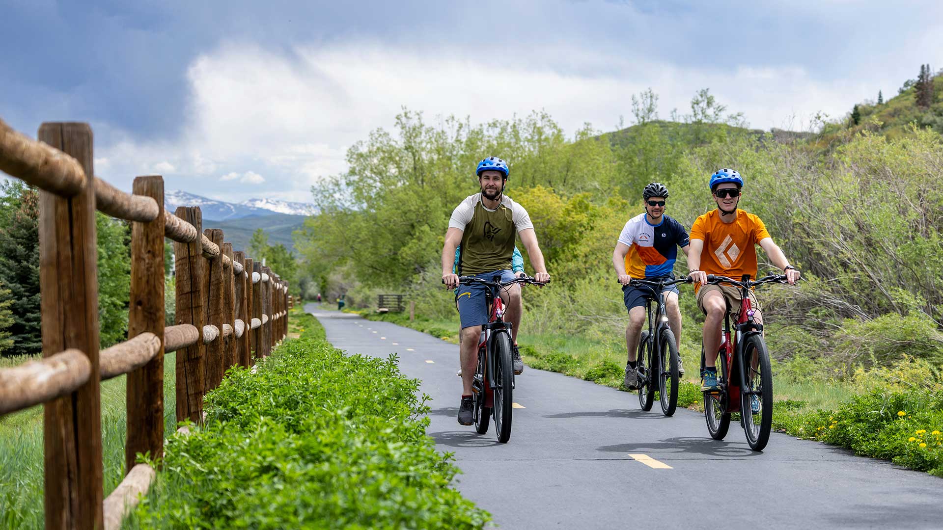 Three cyclists riding on a paved trail with a wooden fence and greenery in the background.