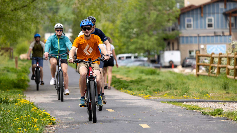 People riding bicycles on a paved path with greenery and buildings in the background