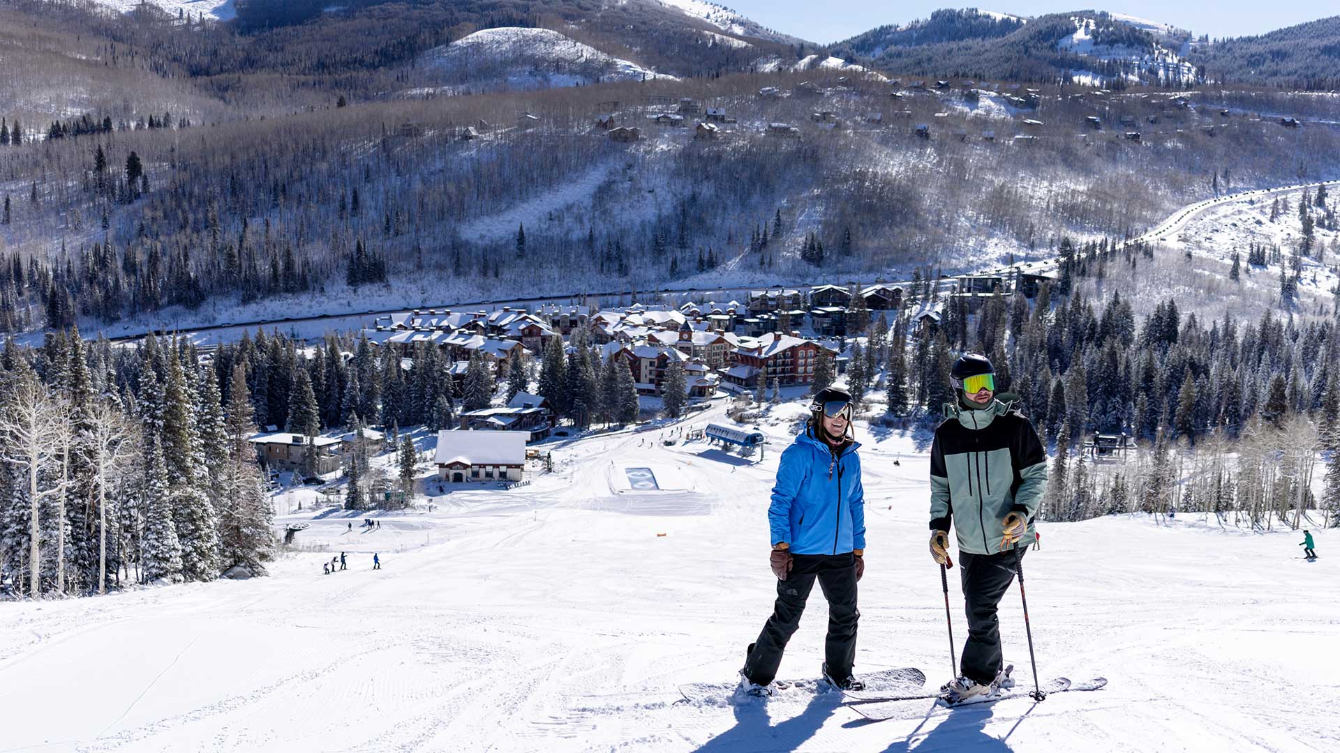 Two skiers standing on a snowy mountain slope with a ski resort village and forested mountains in the background.