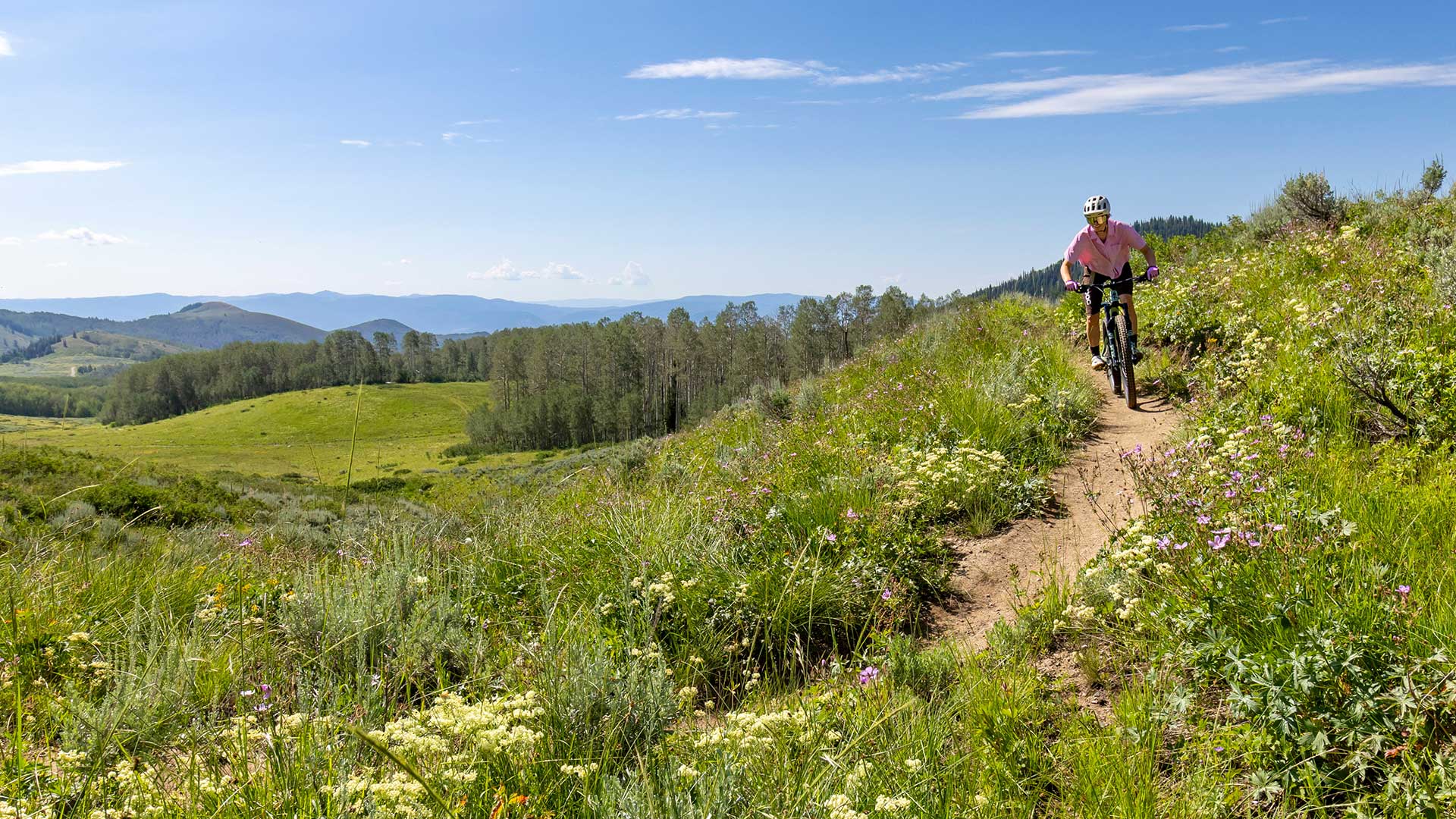 Jans Expert riding a bicycle along a dirt trail on a grassy hillside under a blue sky with distant mountains and trees near Park City, Utah.