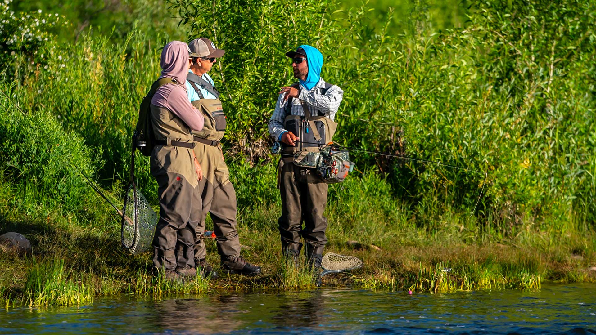 Three people in waders standing by a body of water with greenery in the background