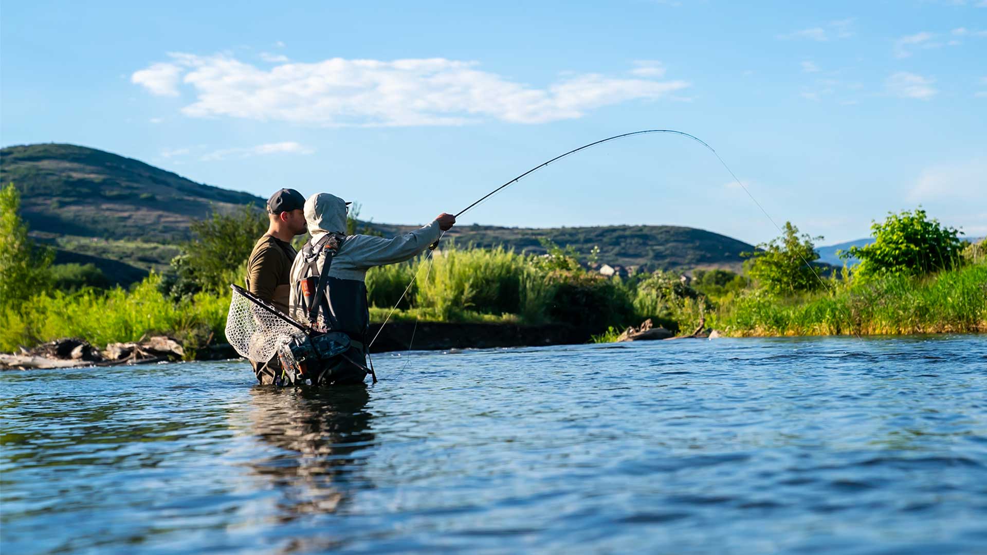 Two people fly fishing in a river with mountains and greenery in the background.