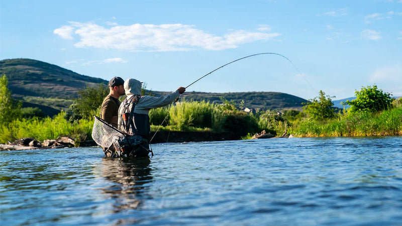 Two people fly fishing in a river with mountains and greenery in the background.