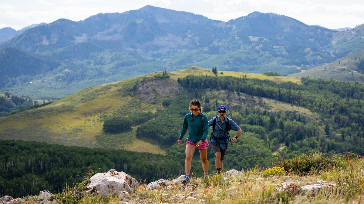Two hikers on a mountain trail with scenic views of mountains and greenery.