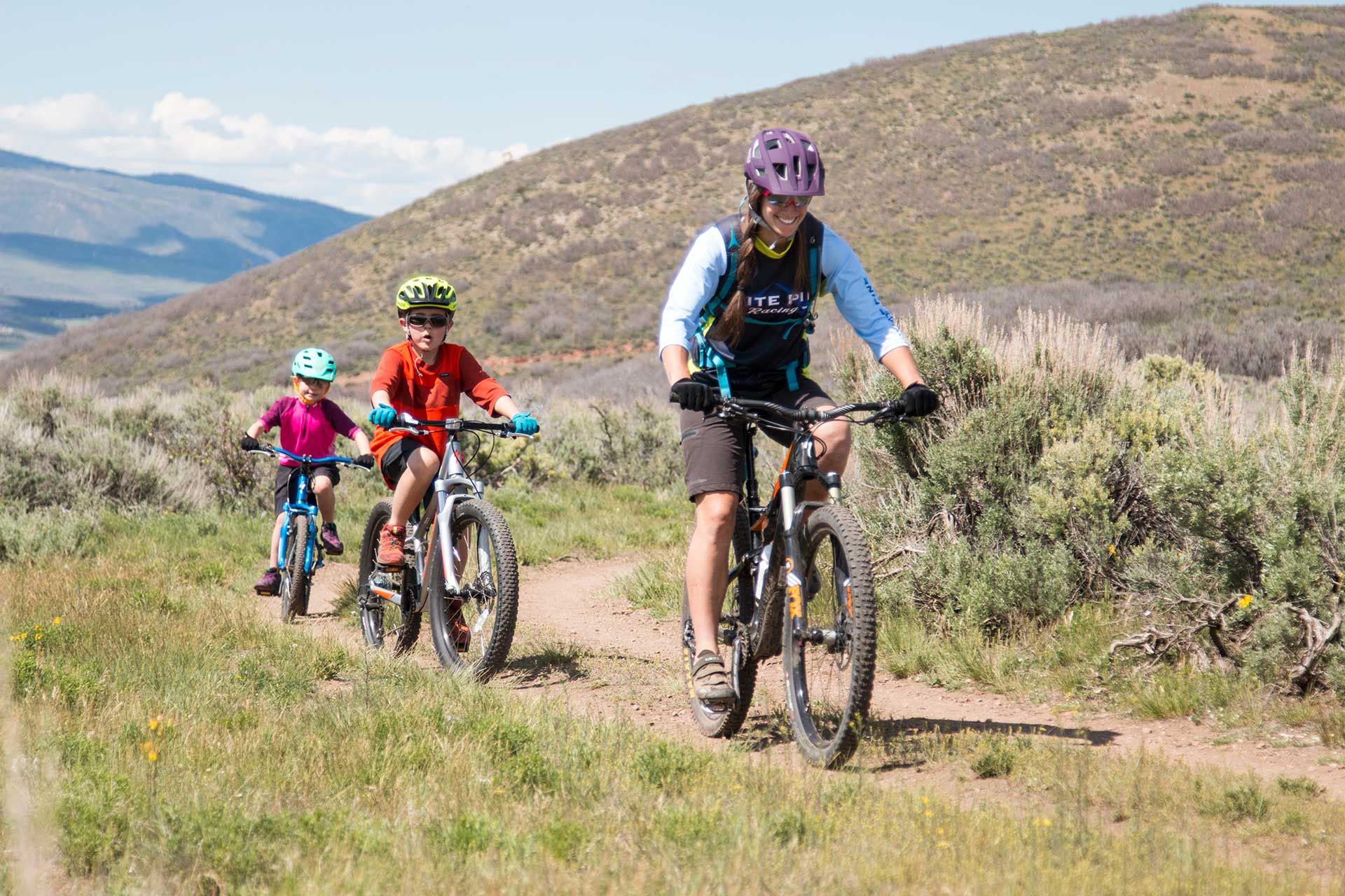 Two children and an adult riding mountain bikes on a trail with mountains in the background