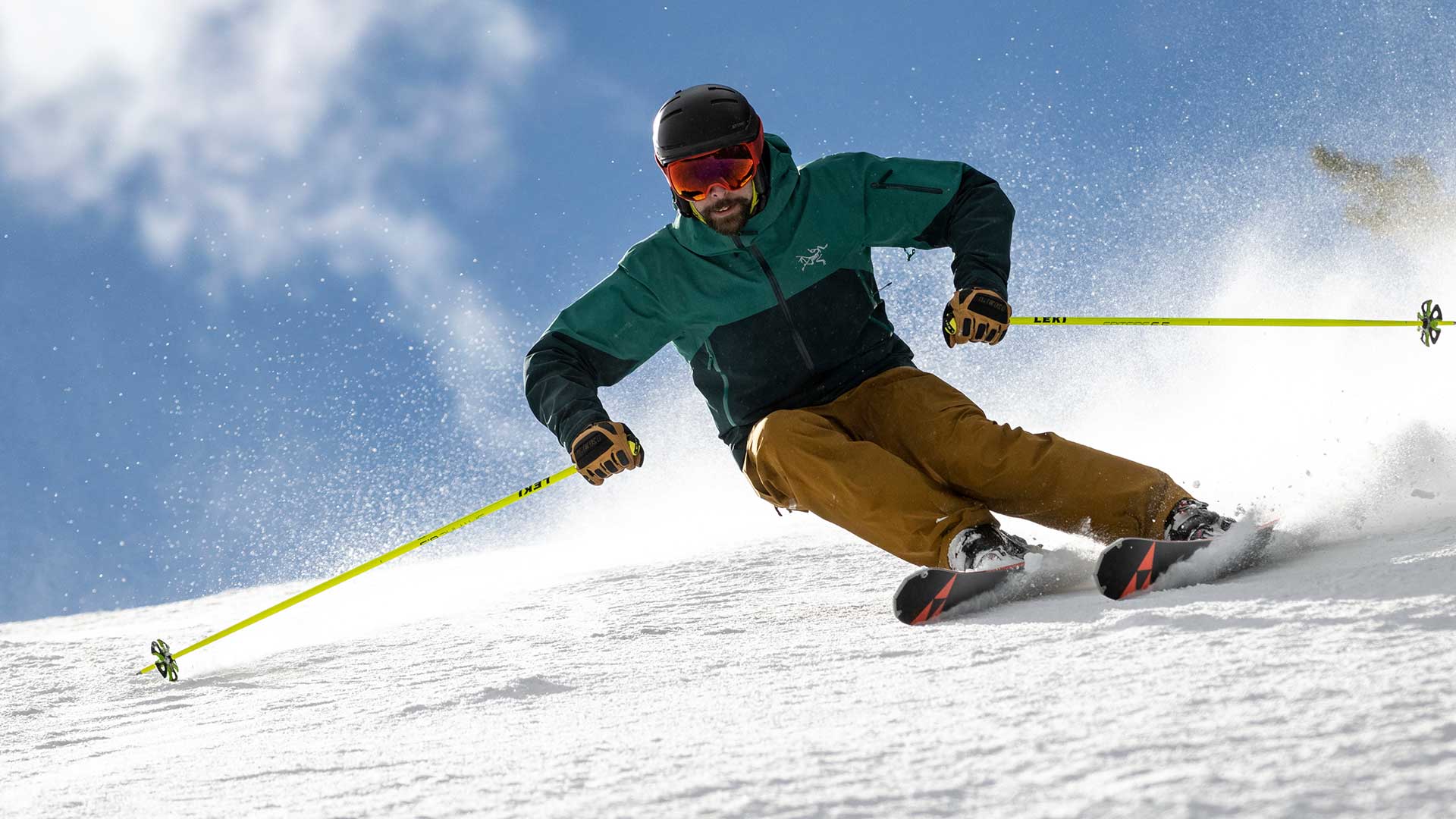 Jans Expert carving through fresh powder on a snowy slope under a bright blue sky, wearing a green Arcteryx jacket and tan pants with yellow ski poles.