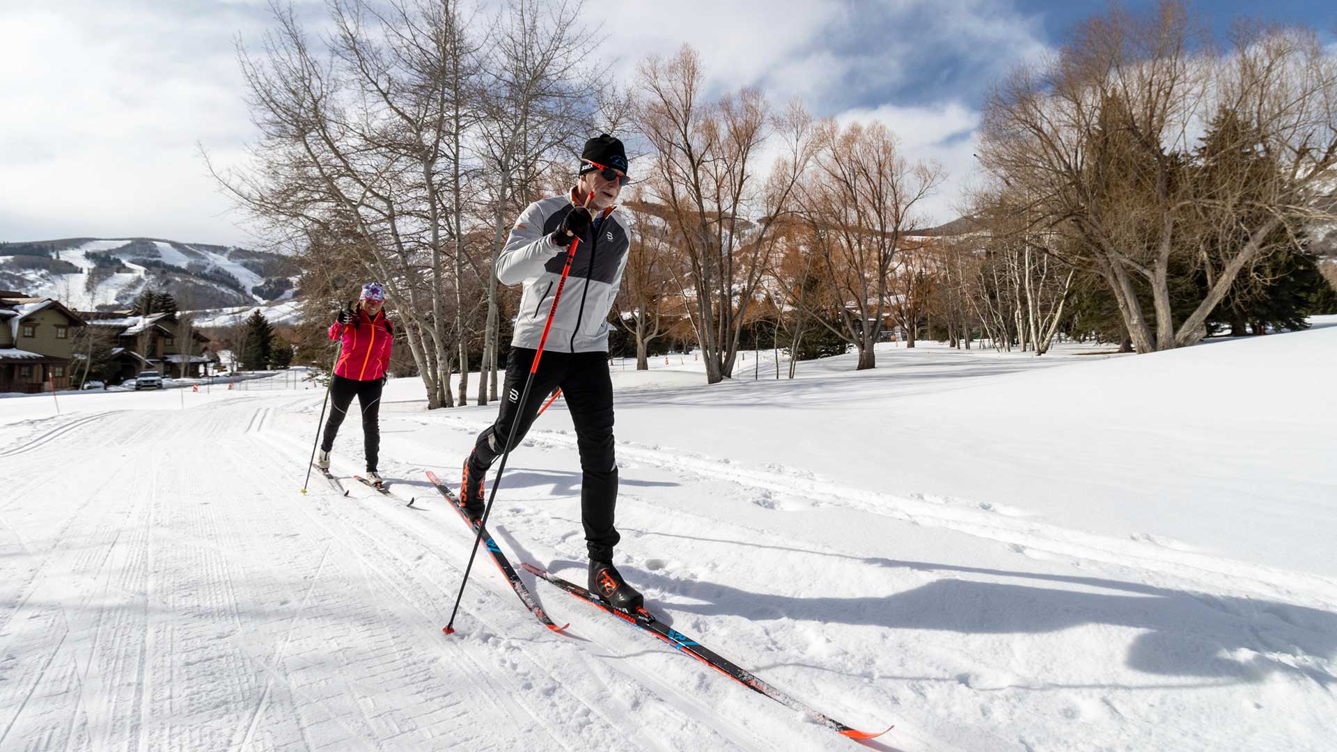 Two people cross-country skiing on a snow-covered trail with a scenic background.