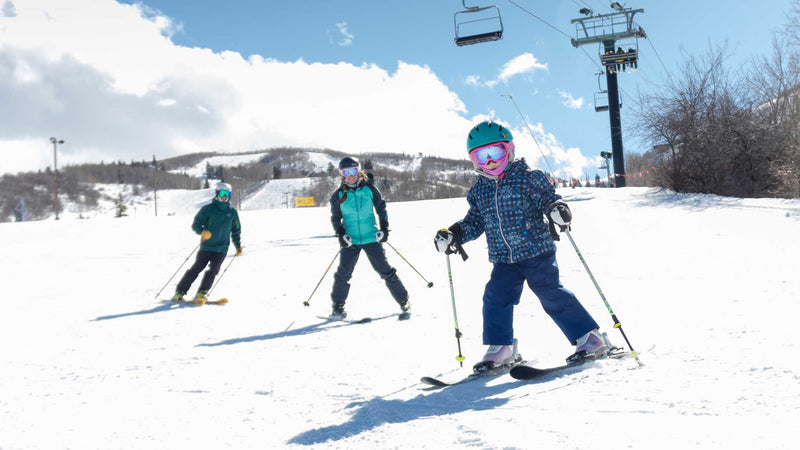 Three skiers on a snowy slope with ski lifts and mountains in the background.