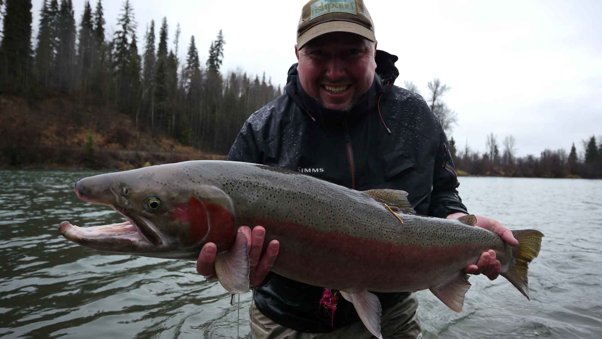 Man holding a large fish by a river with trees in the background
