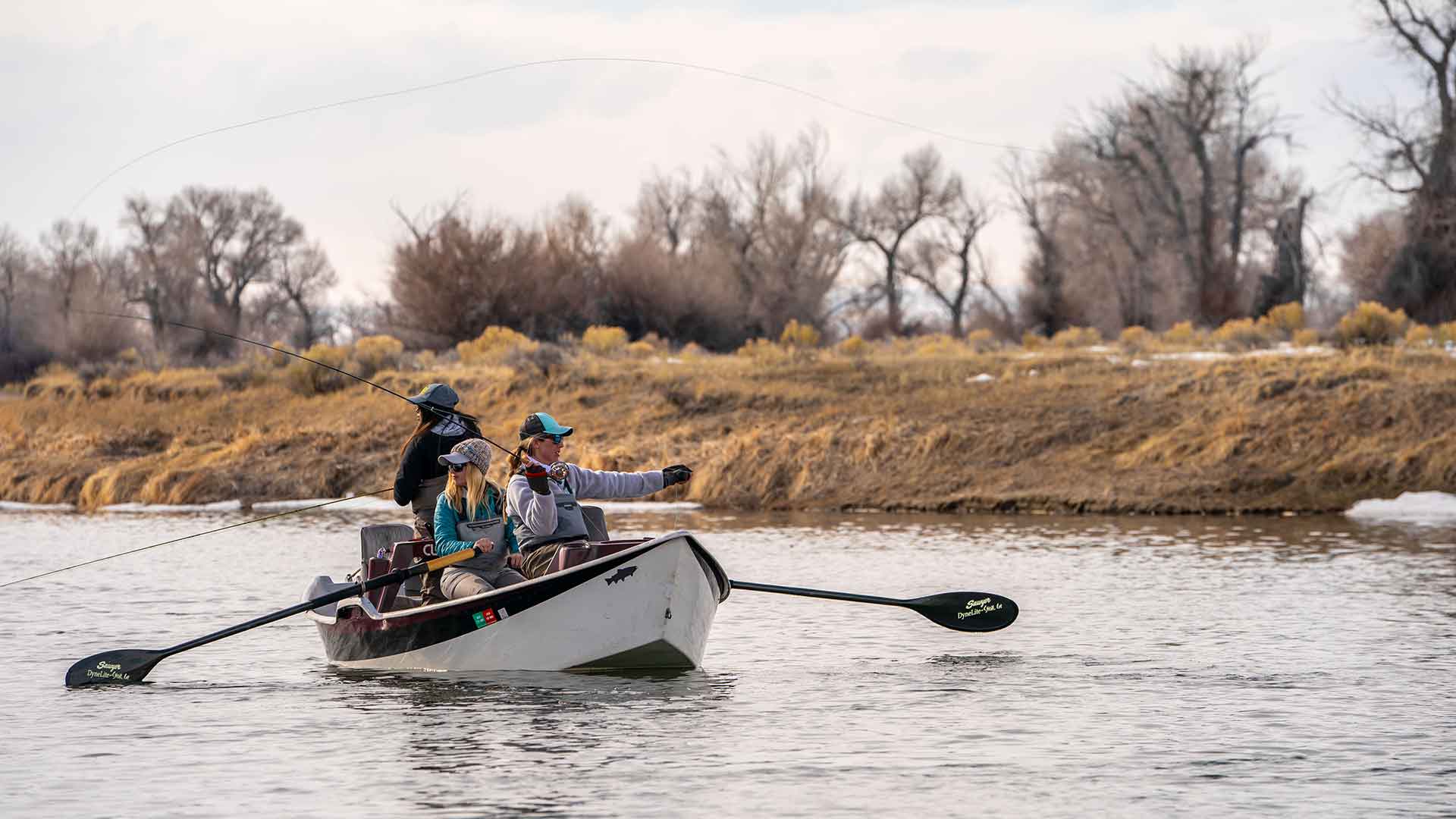 Three people in a small boat on a river with trees and grass in the background