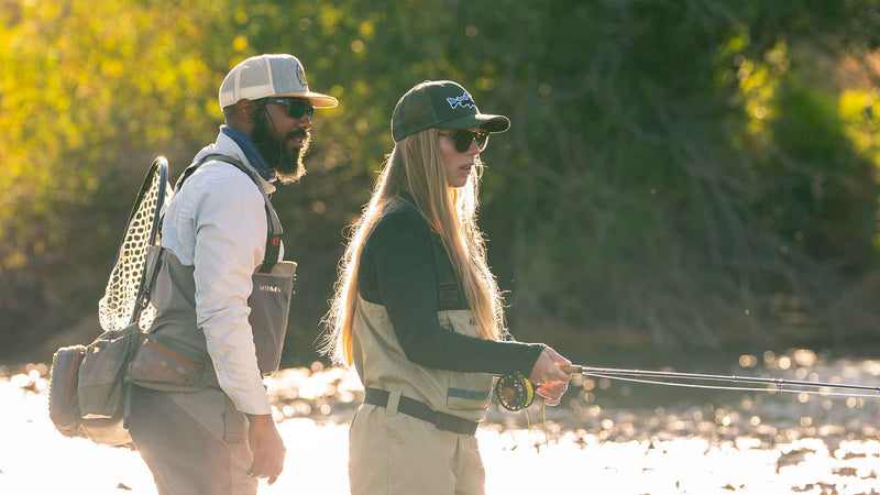 Two people fishing by a river with trees in the background