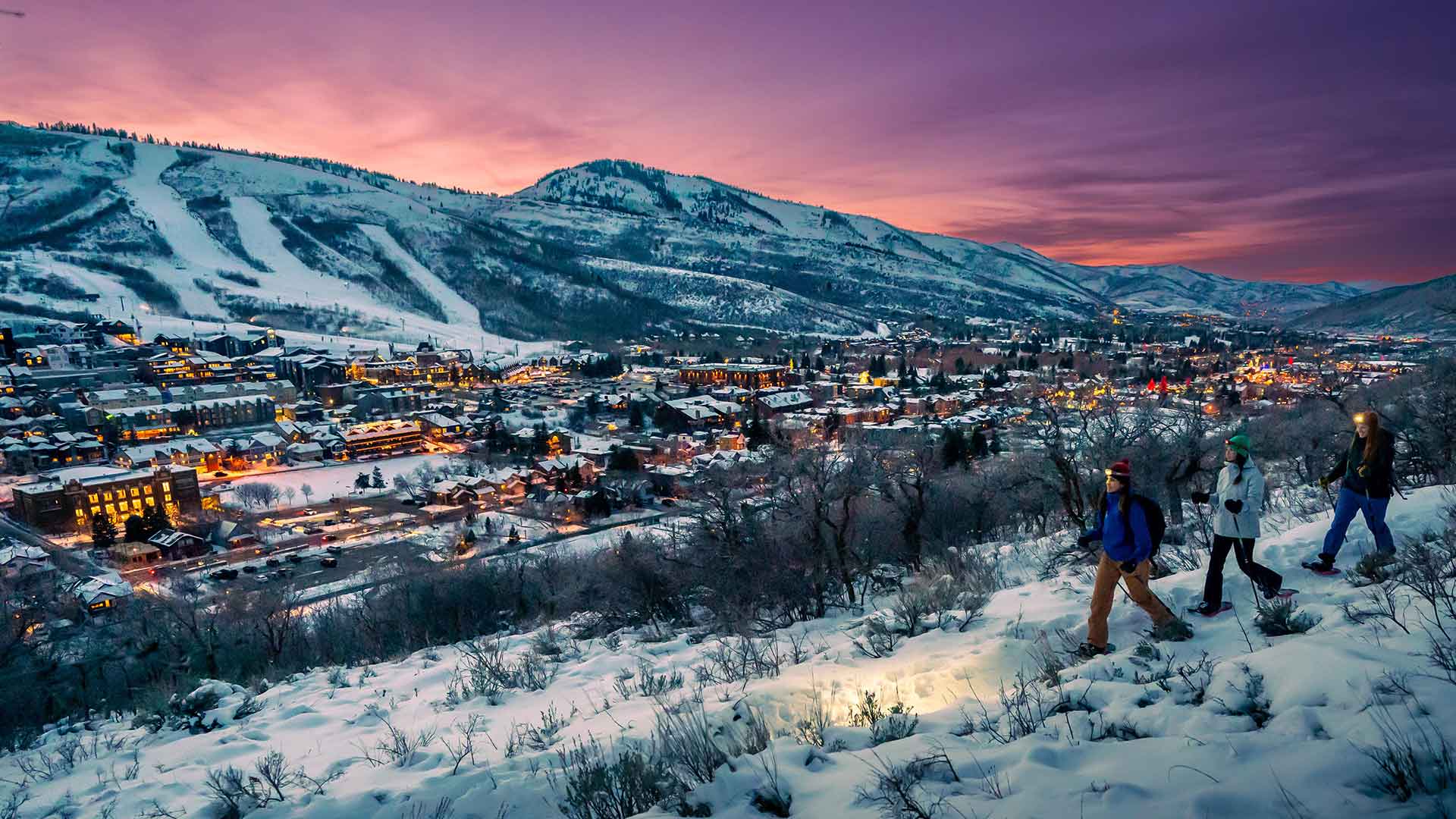 Three people with skis and snowboards standing on a snowy hill overlooking a ski resort at dusk.