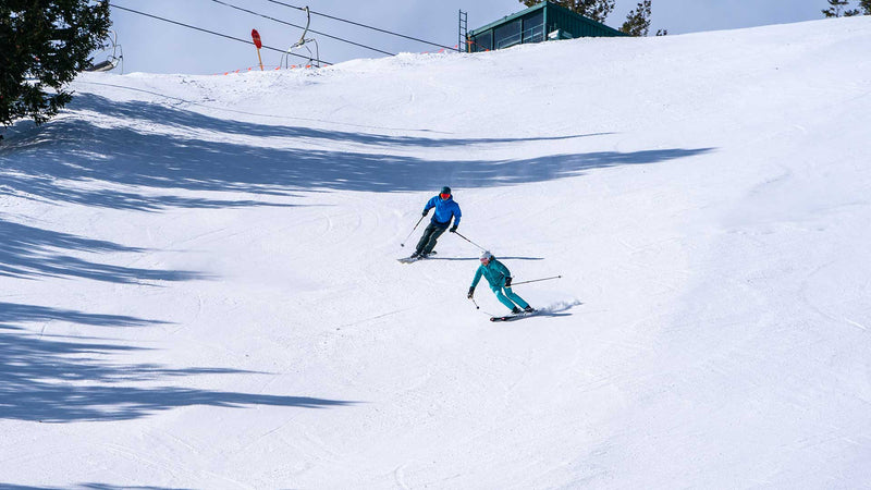 Two skiers on a snowy slope with trees and ski lift in the background.