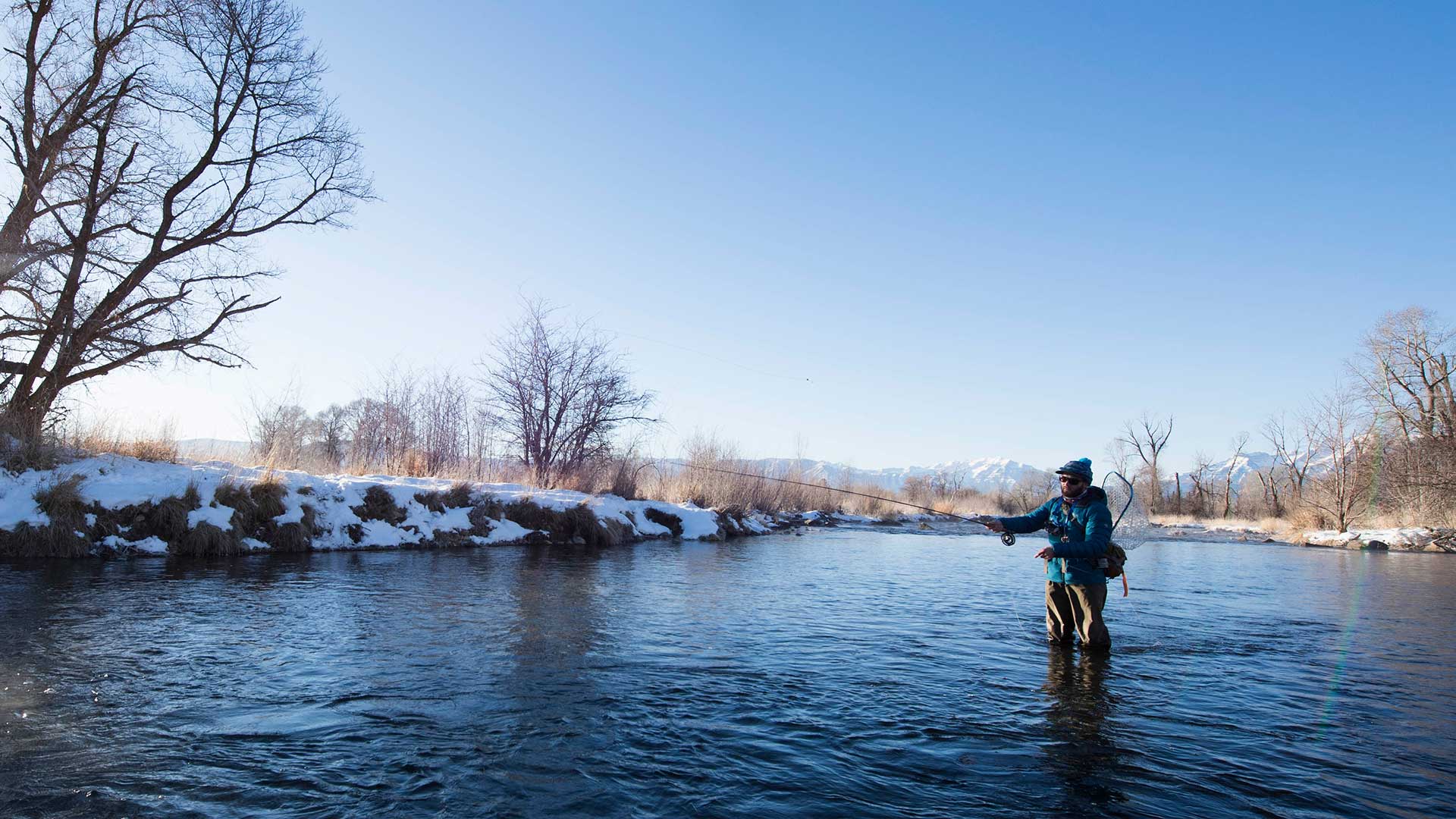 Person fly fishing in a river with snow-covered banks and trees.