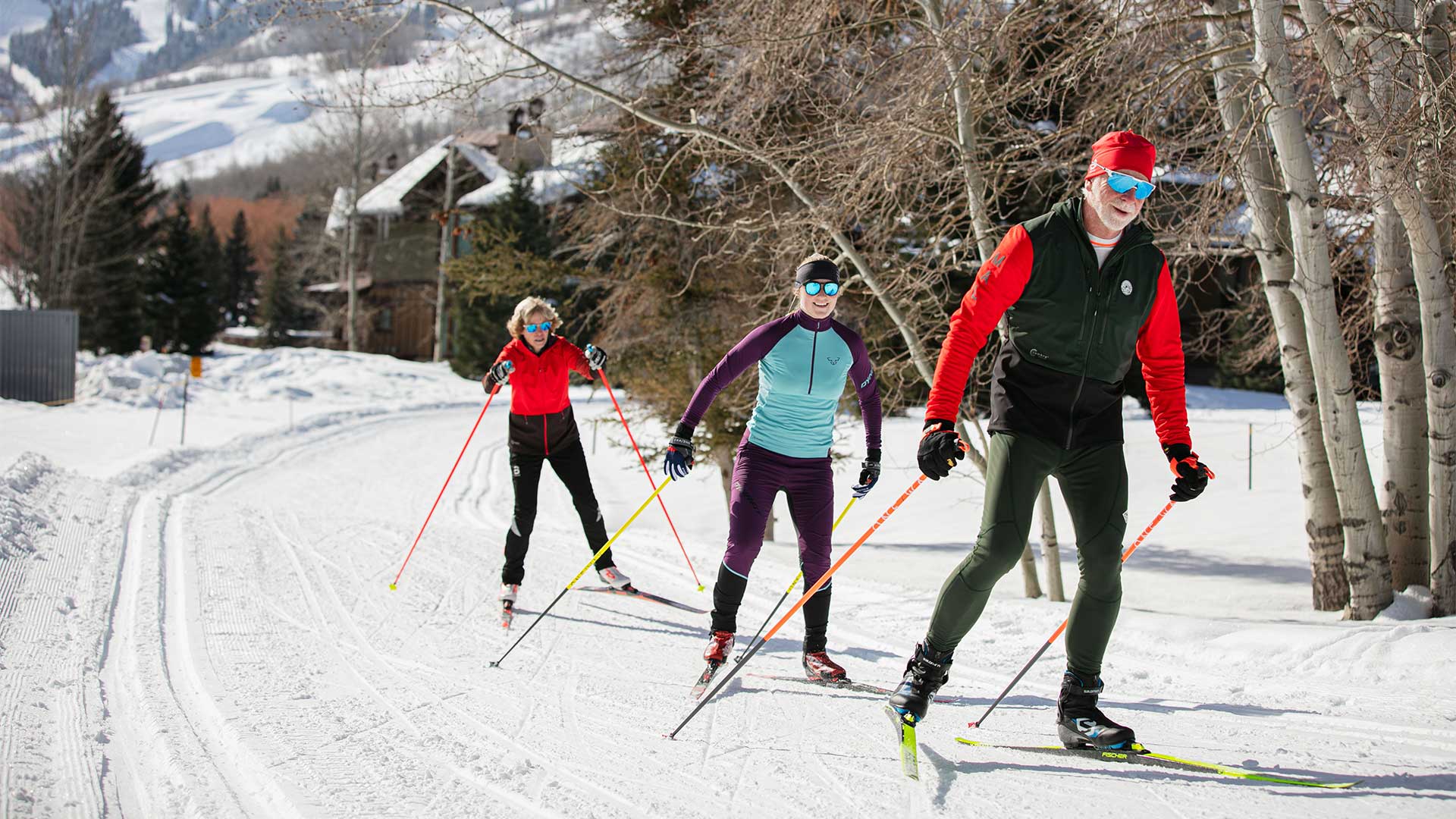 Three cross-country skiers on a snow-covered trail with ski tracks and trees in the background.