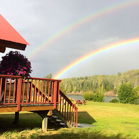Double rainbow over a cabin with a deck and flower pot, surrounded by trees and water.