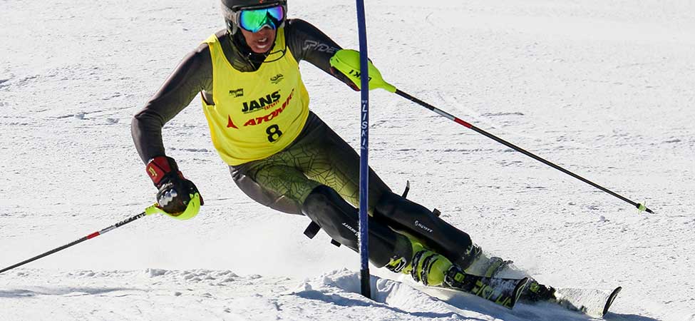 A skier in a yellow and black racing suit with the JANS brand and number 8, wearing goggles and holding ski poles, carving through the snow.