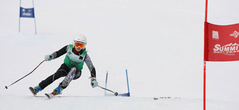 A skier in a green and black racing suit leans into a turn on a snowy slope, with a red Summit ski gate flag visible on the right.