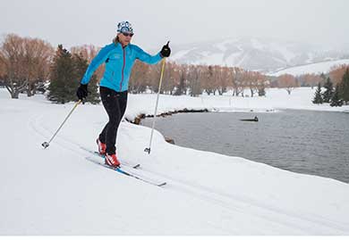 Person cross-country skiing on a snowy landscape with a lake and mountains in the background.