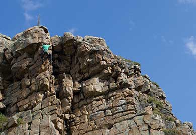 Person climbing a rocky mountain peak with a clear blue sky in the background
