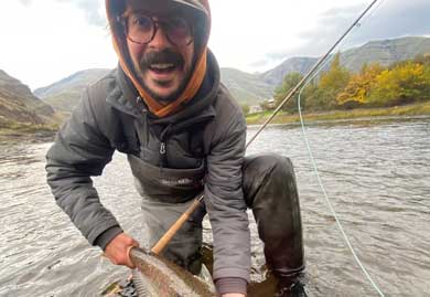 Man holding a fish while fishing in a river with mountains in the background