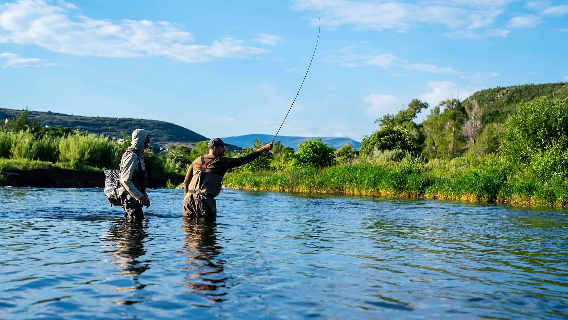 Two people fly fishing in a river with greenery and mountains in the background.