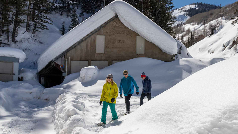 Three people walking through deep snow towards a snow-covered building with 'Village Gym' sign.