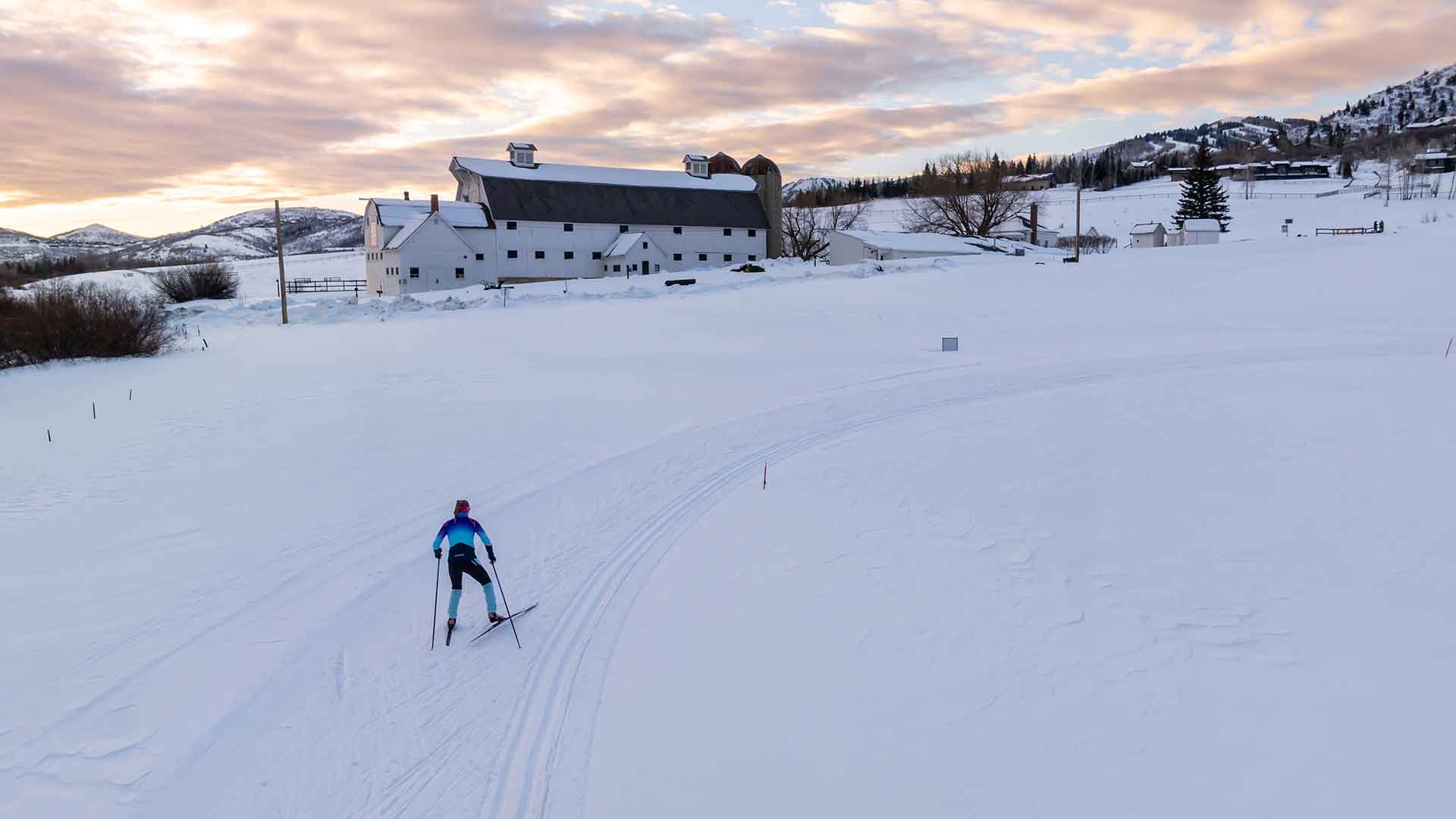 Person cross-country skiing on a snow-covered field with a building and mountains in the background.