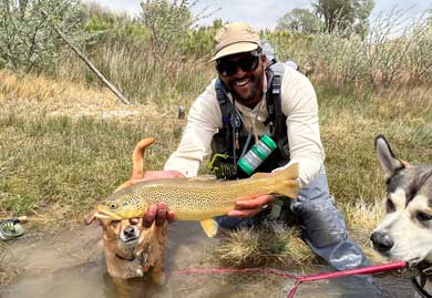 Man holding a fish by a river with two dogs by his side