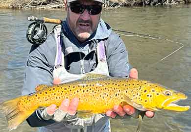 Man holding a large fish by a river with fishing equipment.